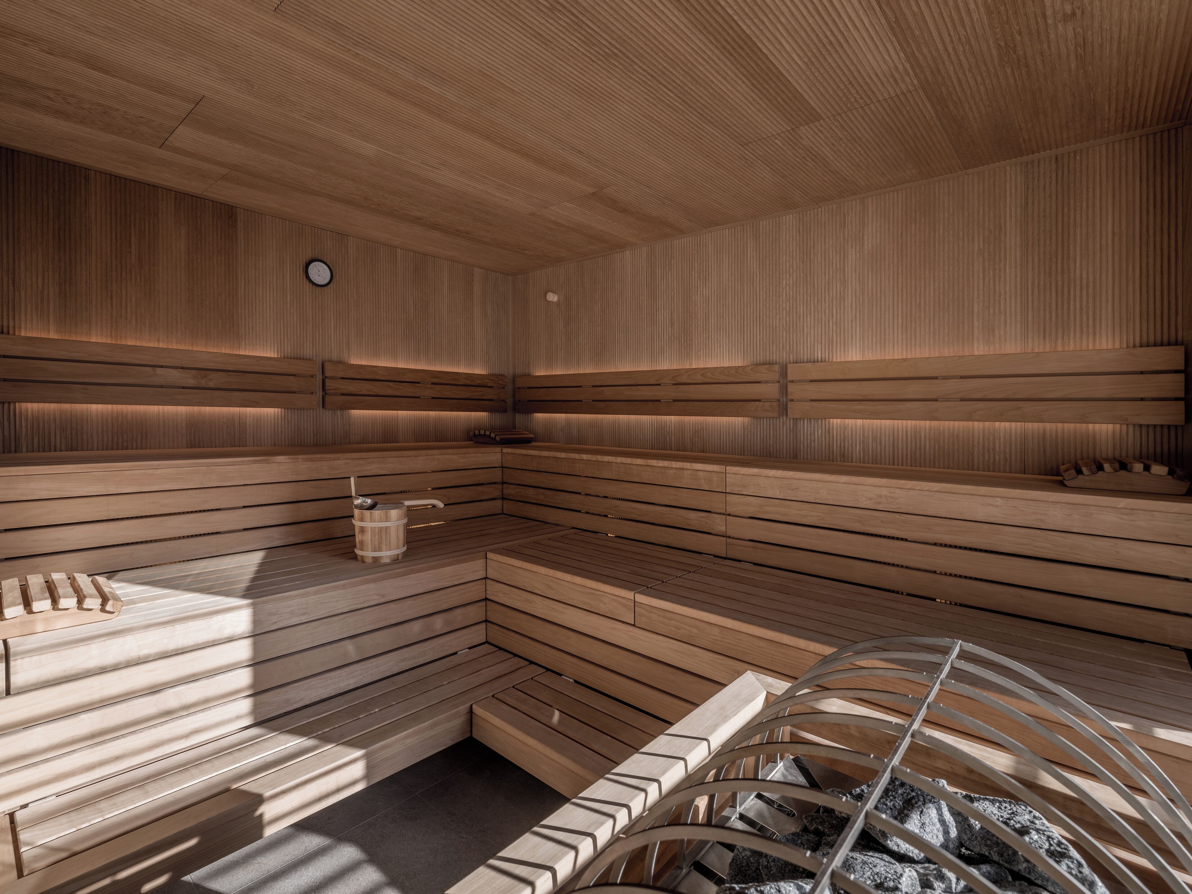 A sauna with wooden benches and a stone bench, a wooden wall with a clock, and a metal bucket.