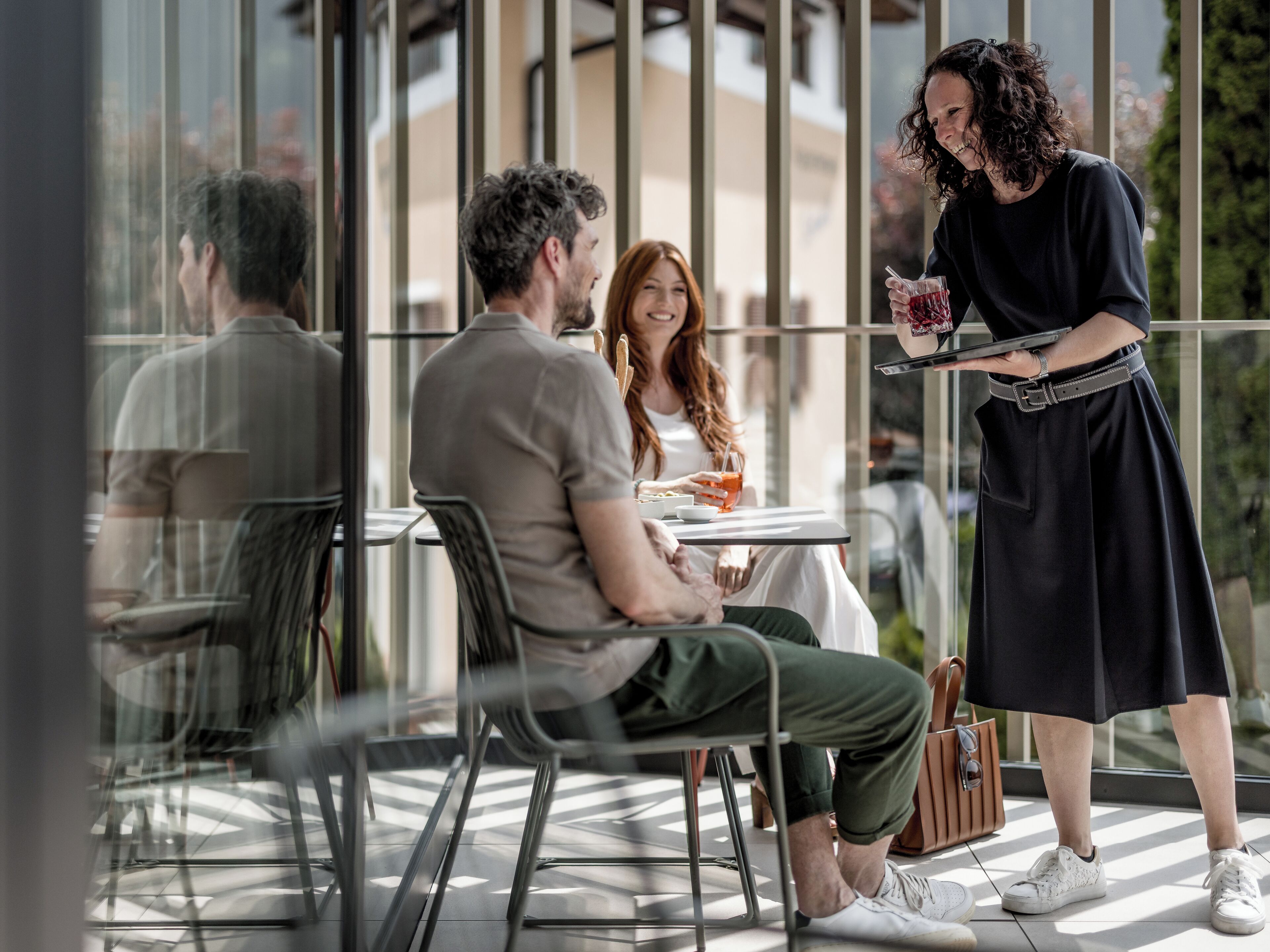 Un espacio al aire libre moderno con separador de vidrio, un camarero sirviendo una bebida, y una mesa con una computadora portátil.