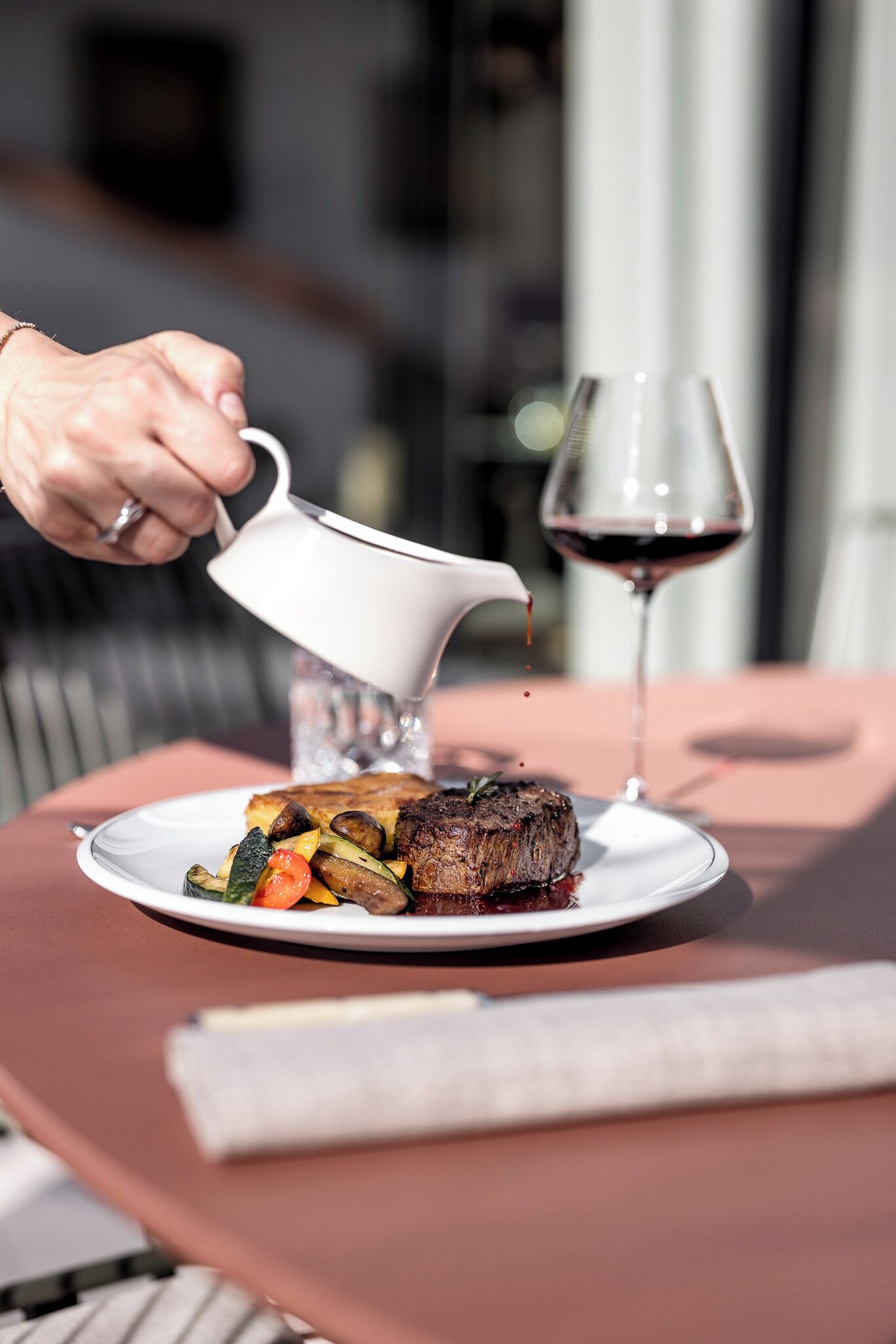 A steak with vegetables on a plate, a wine glass, and a person pouring a drink.