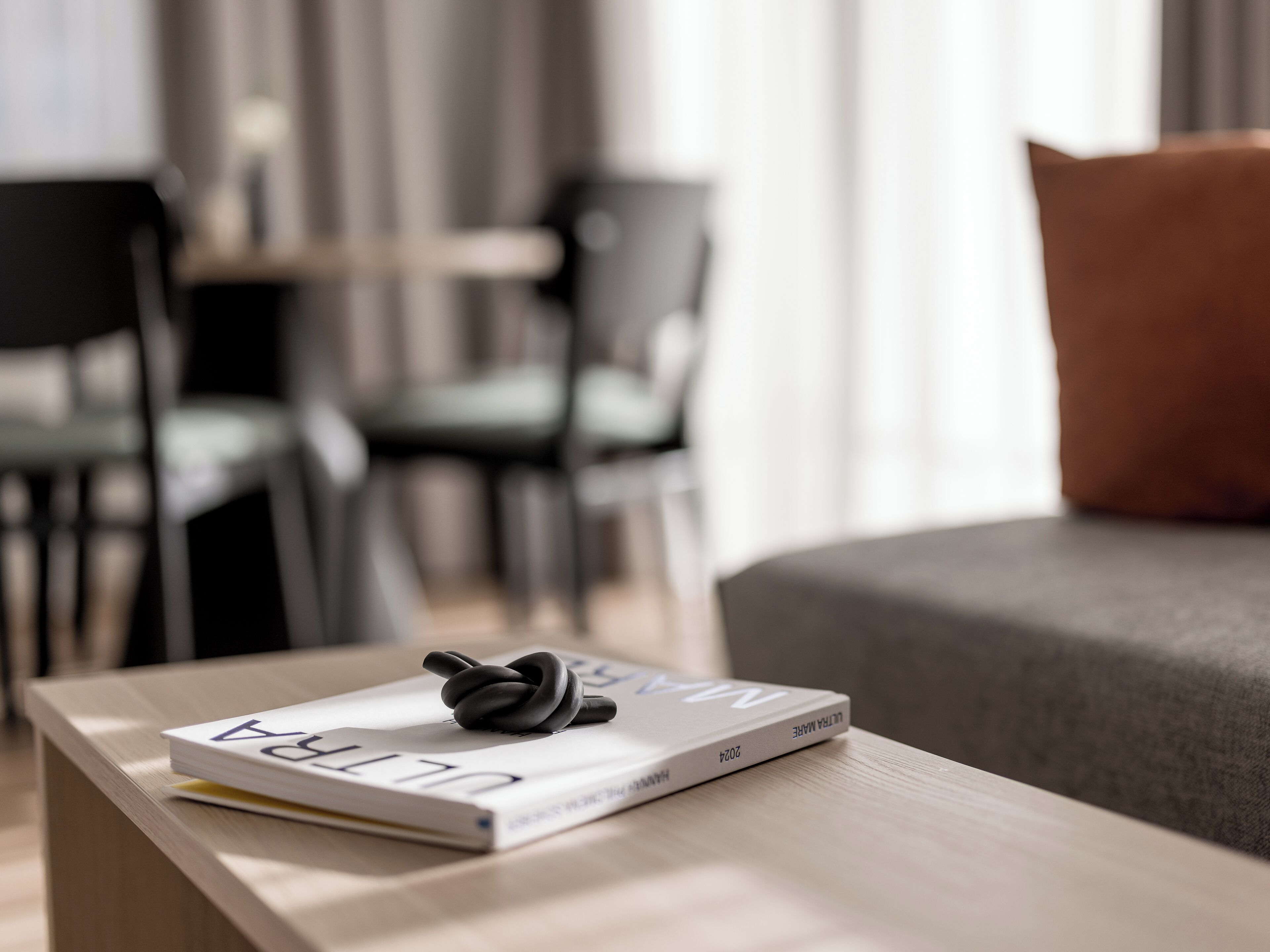 A neatly arranged room with a modern design, featuring a sofa, a coffee table with a book and a pair of earphones, and a dining area with chairs and a table.