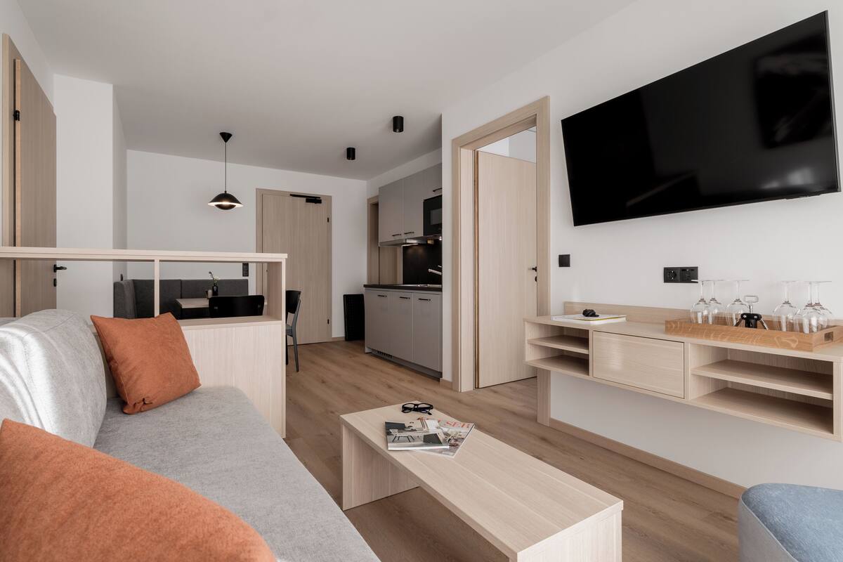 A modern, well-lit living room with a grey sofa, orange cushions, a wooden coffee table, and a TV mounted on the wall.