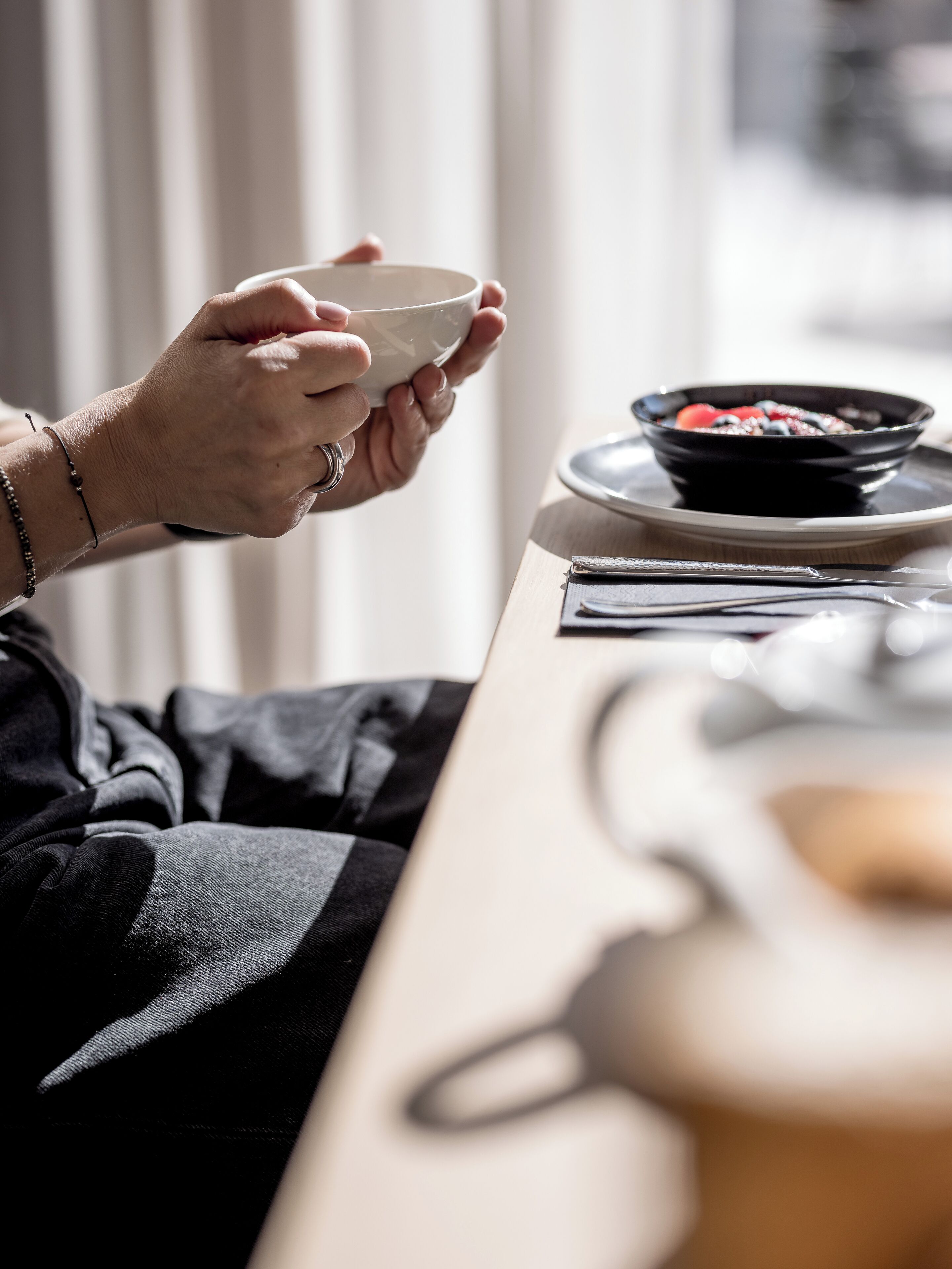 A person holding a white cup, with a bowl of food and a plate on a table.