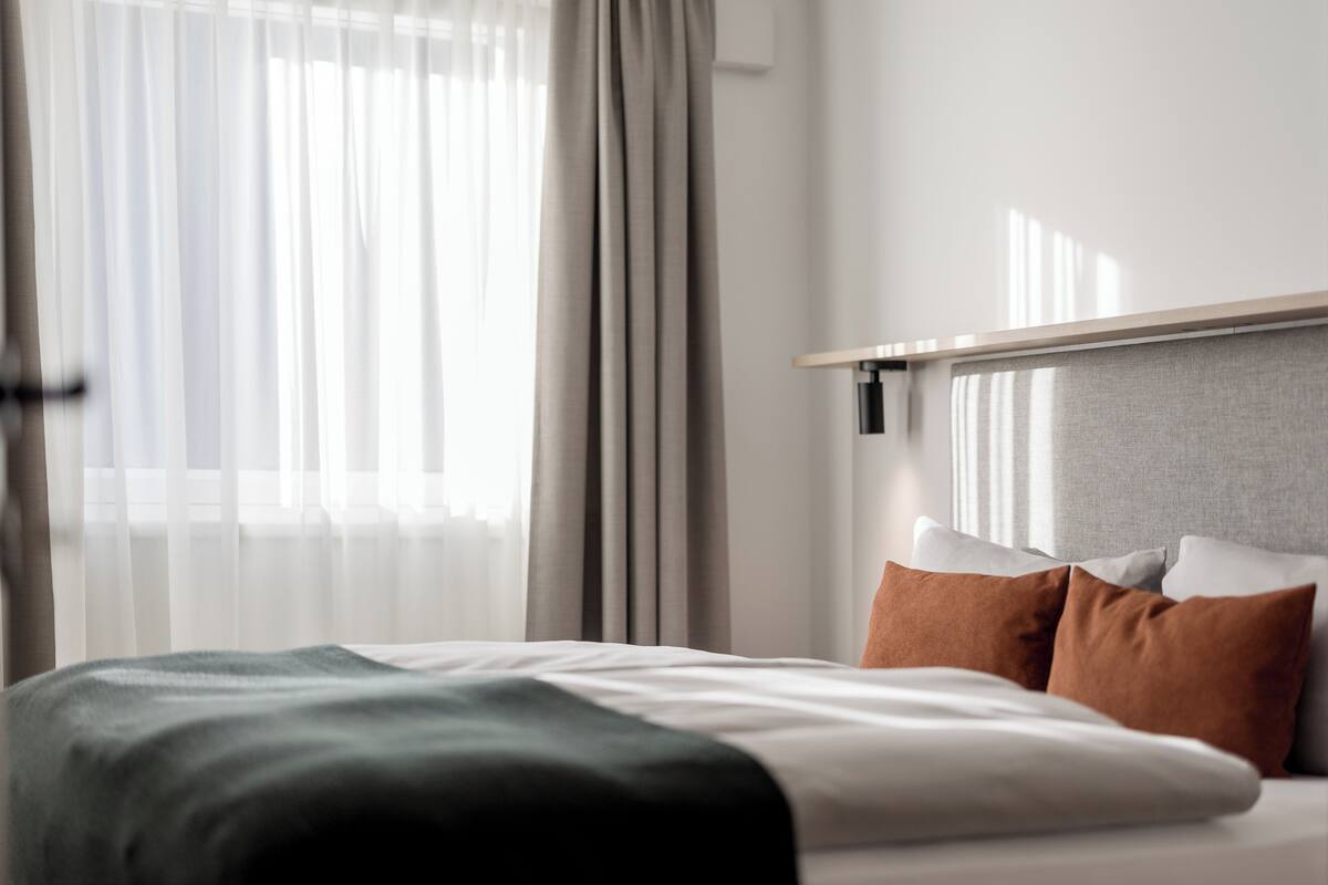 A modern hotel room with a neatly made bed, a headboard, and a large window with sheer curtains.