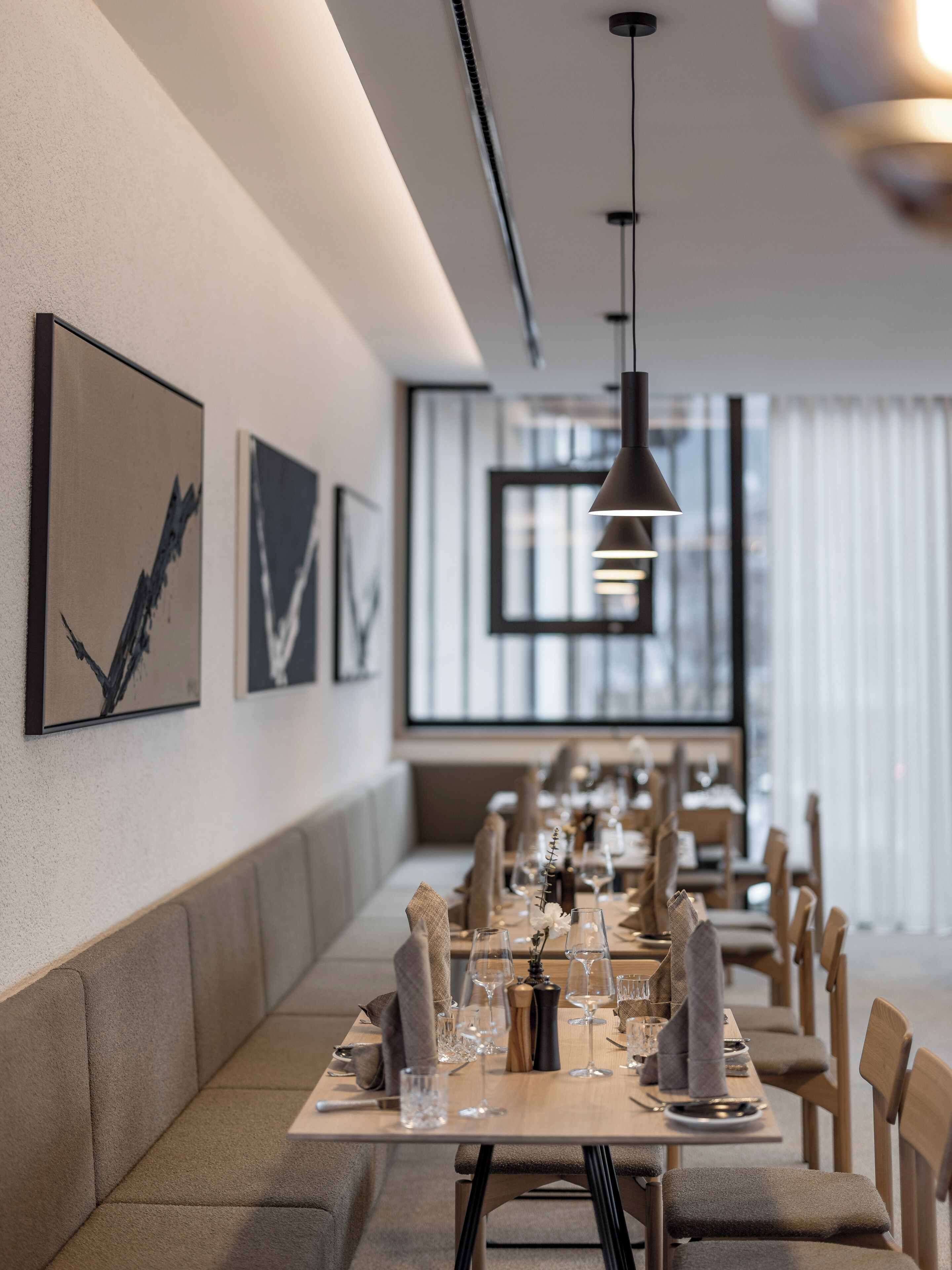 A modern dining area with a long table set for a meal, framed artwork on the walls, and natural light from large windows.