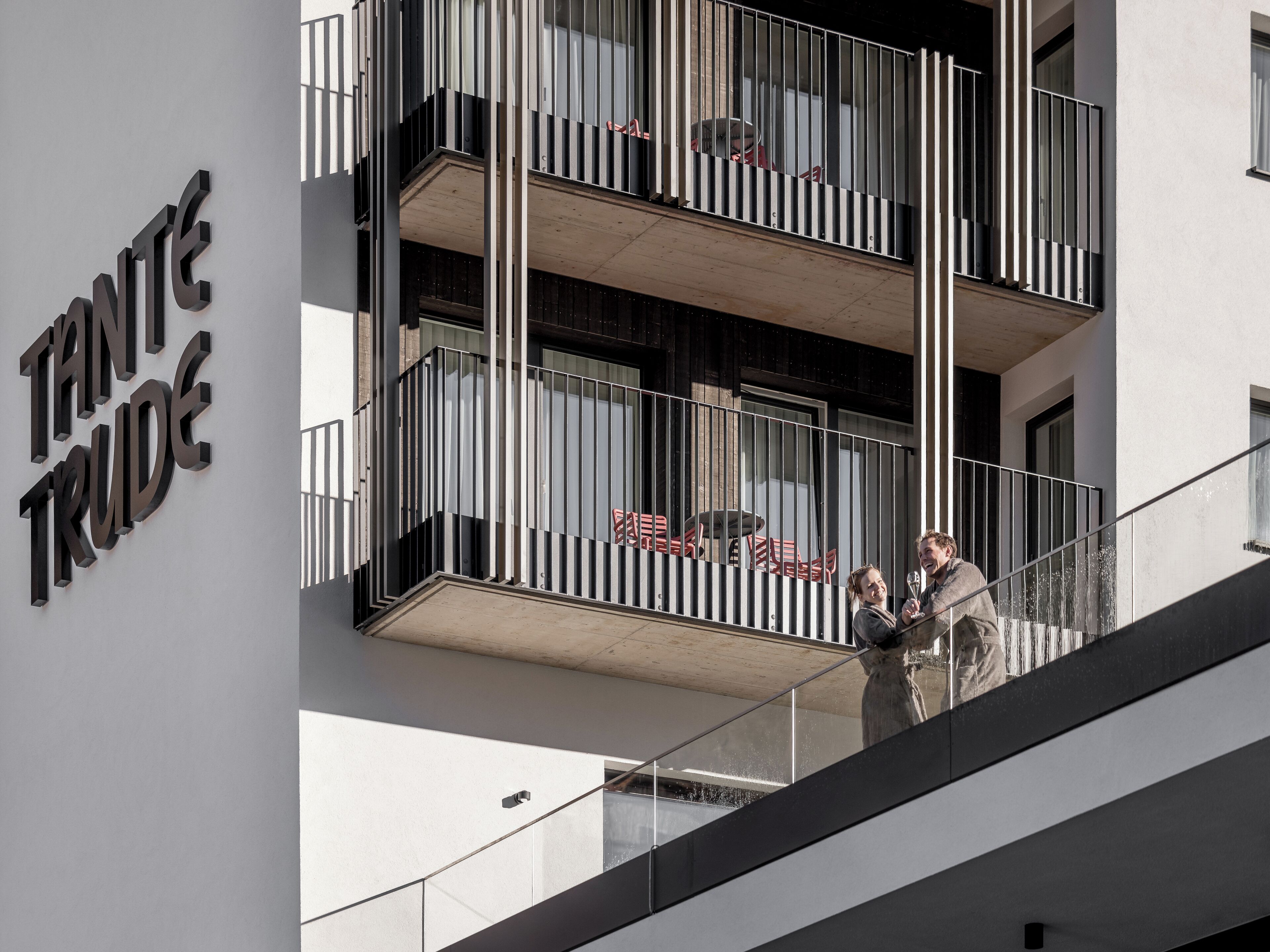 A modern building with a sign reading "TANTE TRUDE". It features a balcony with metal railings and a view of the street below.