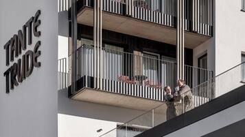 A modern building with a sign reading "TANTE TRUDE". It features a balcony with metal railings and a view of the street below.