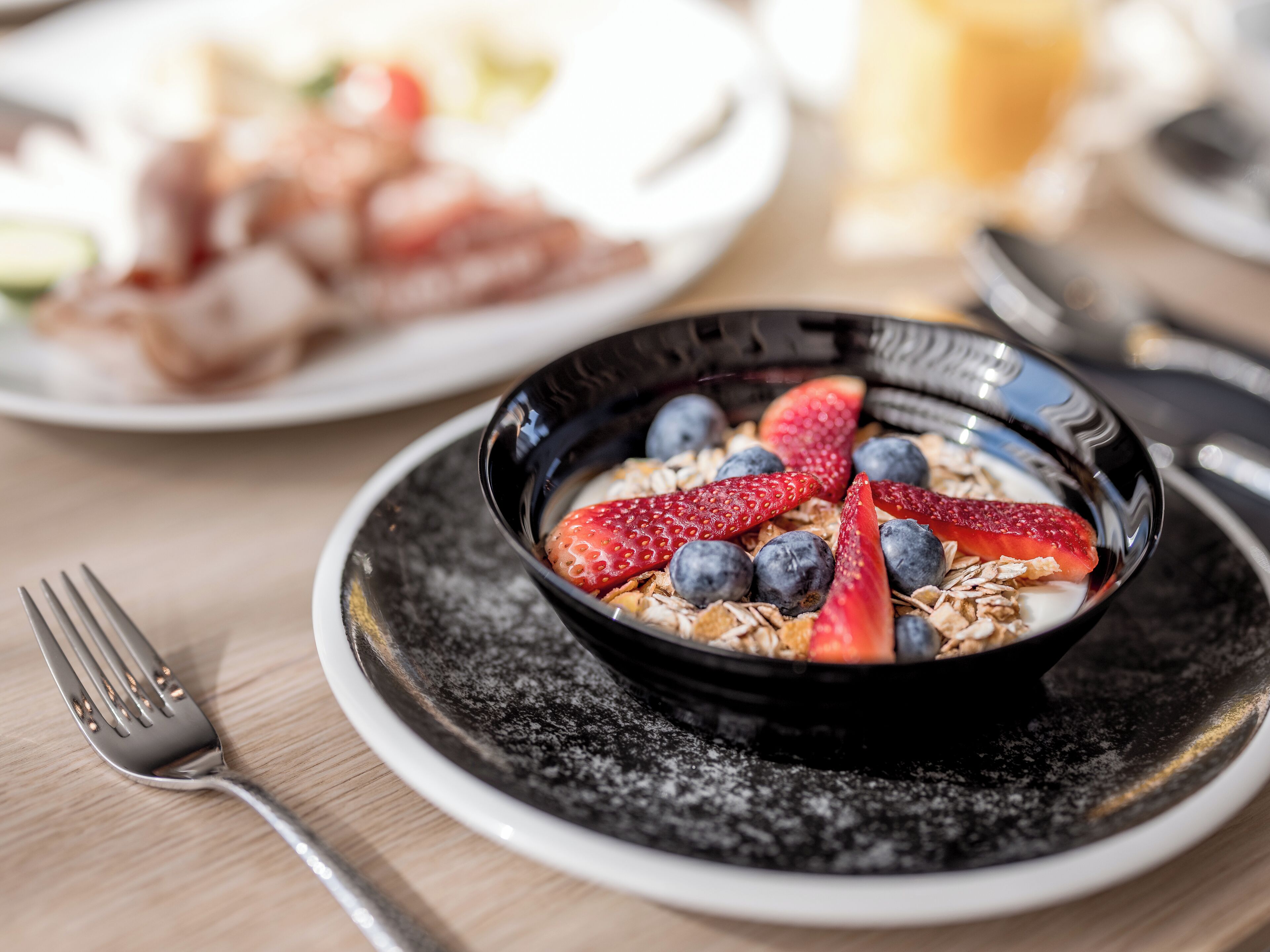 Desayuno con un tazón de yogur cubierto de fresas y arándanos, un tenedor y un plato de tocino.