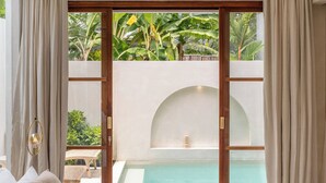 A bedroom with a large bed, a view of a swimming pool, and a door leading to an outdoor area with greenery. - Villa Inka (Canggu)