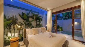 A bedroom with a large bed, a nightstand, a potted plant, and a view of a pool and garden through a glass door. - Villa Inka (Canggu)