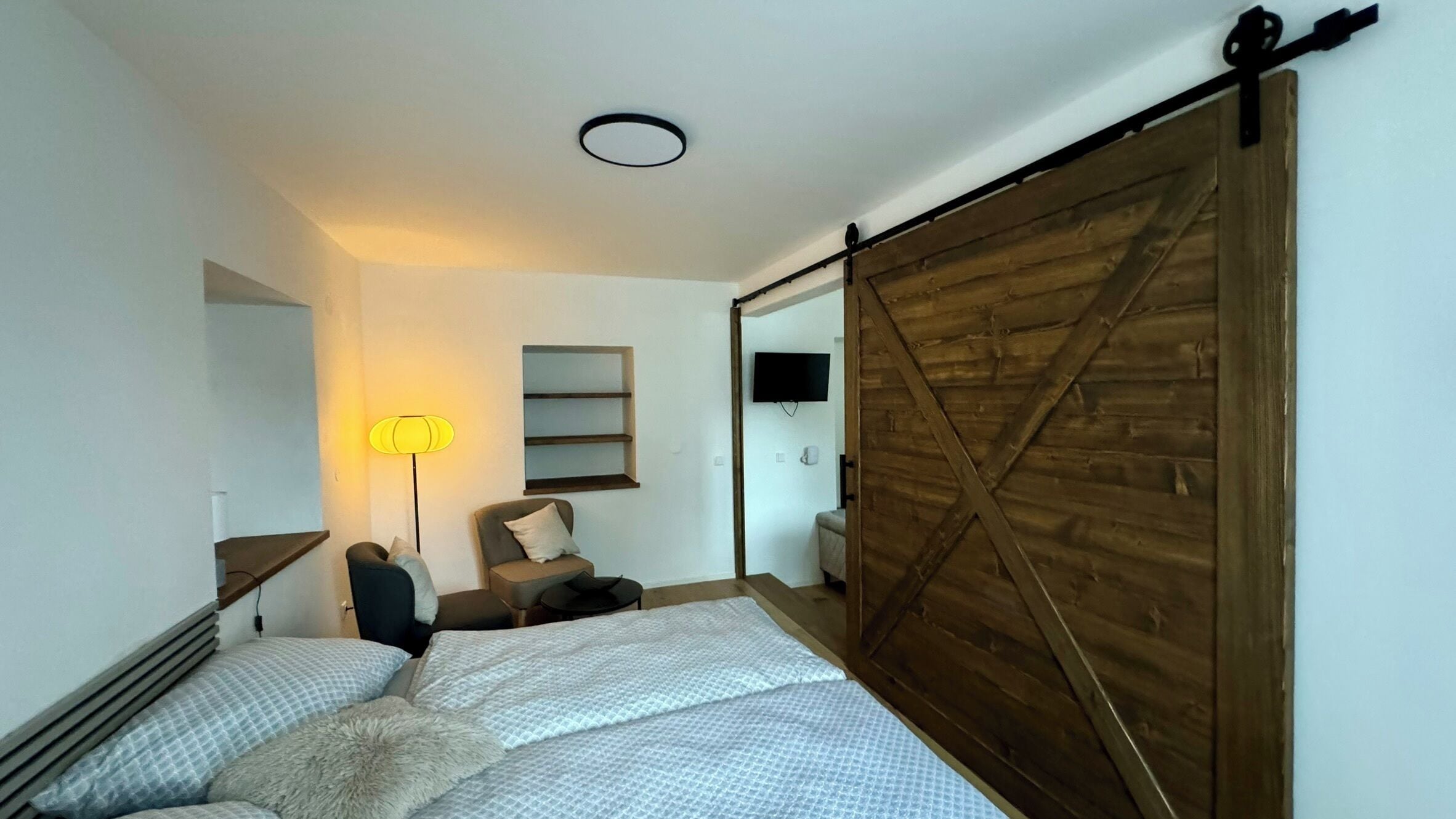 A bedroom with a bed, a chair, a desk, a lamp, and a TV.