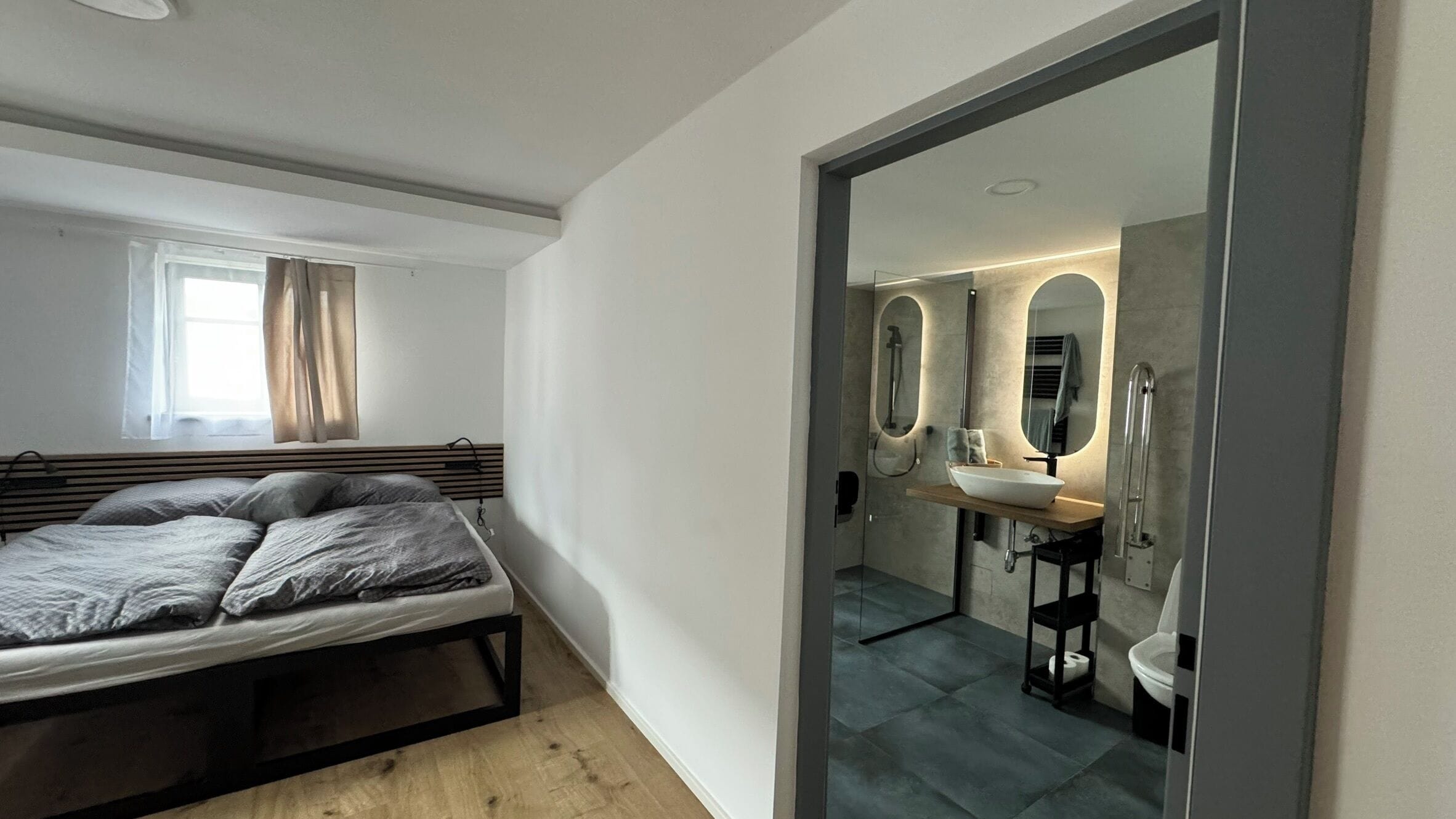 Un dormitorio con cama, ventana con cortinas y baño con lavamanos, espejo y ducha.