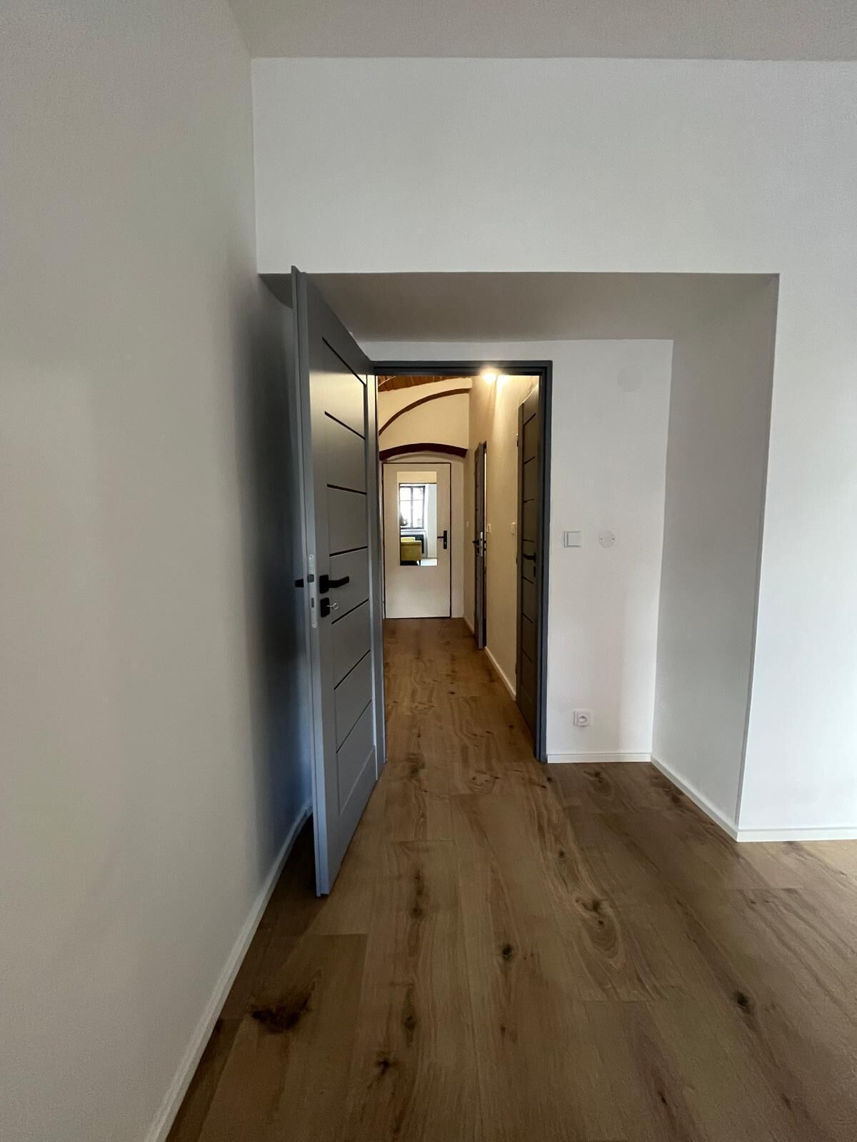 Un couloir au sol en bois et aux murs blancs, qui mène à une pièce dotée d’une porte.
