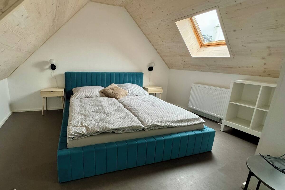 A bedroom with a sloped ceiling, a bed with a teal headboard, a small bedside table, a radiator, and a window.