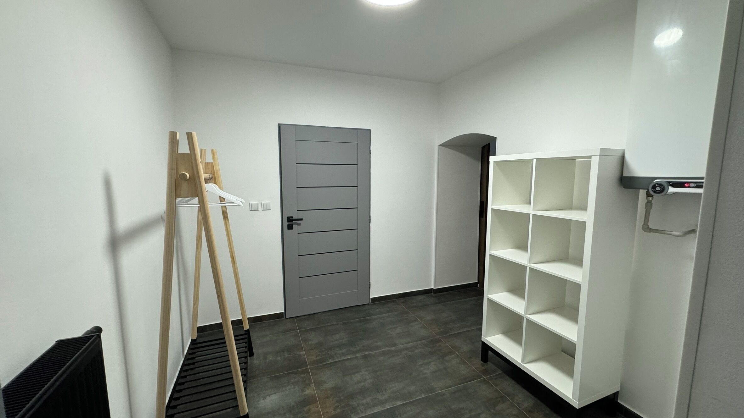 Un passadís amb una porta gris, un moble d'estanteries blanc i una escala de fusta adossada a la paret.