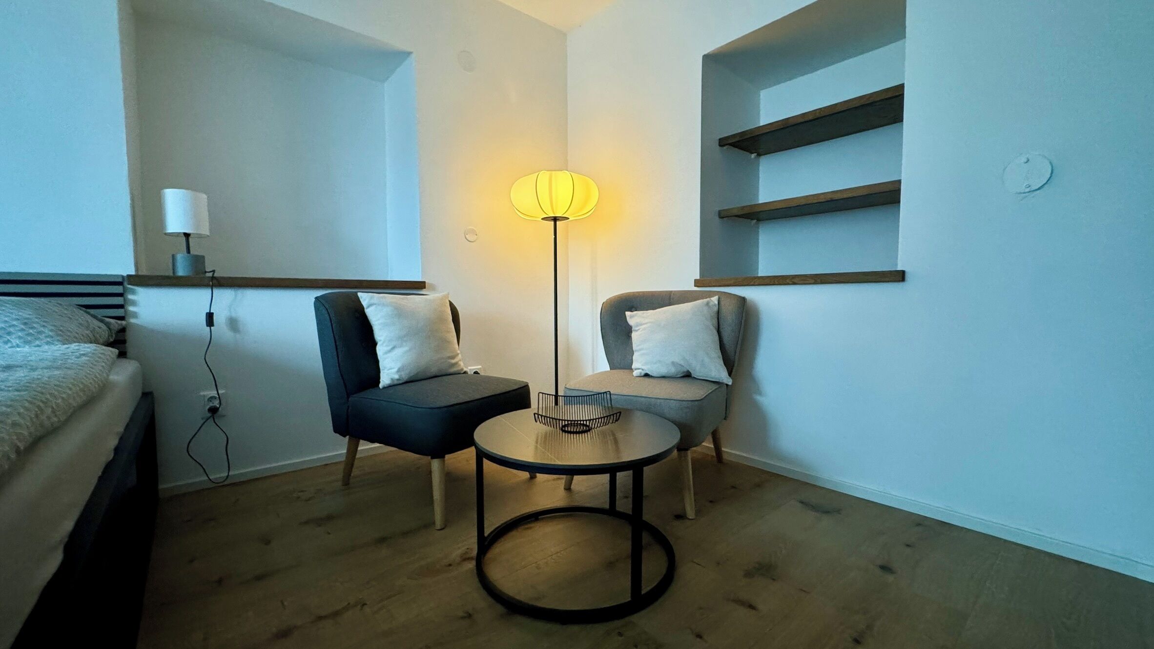 Una habitación pequeña y moderna con una cama, dos sillas, una mesita y una lámpara de pie.