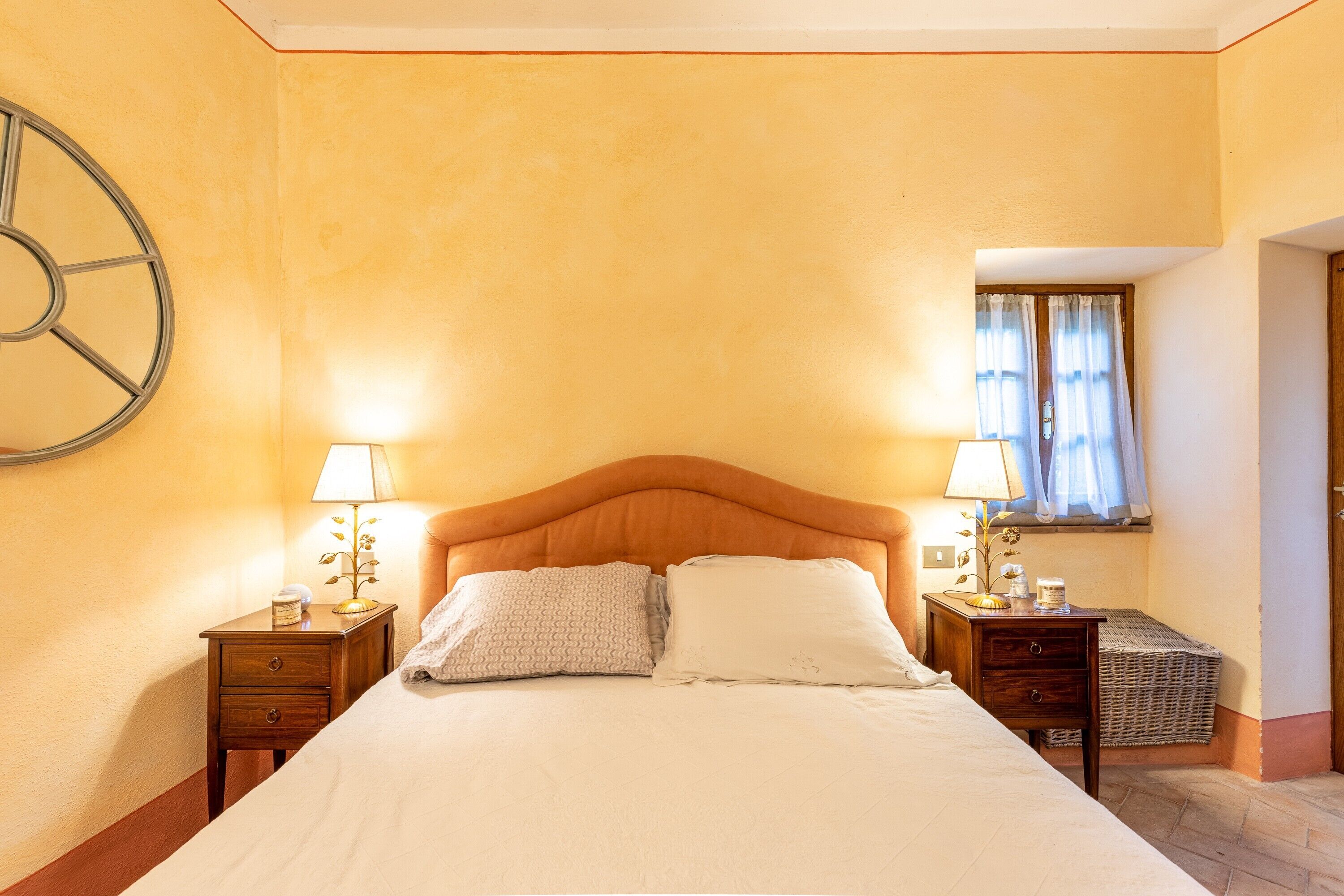 A hotel room with a bed, two bedside tables, a lamp, a clock on the wall, and a window with curtains.