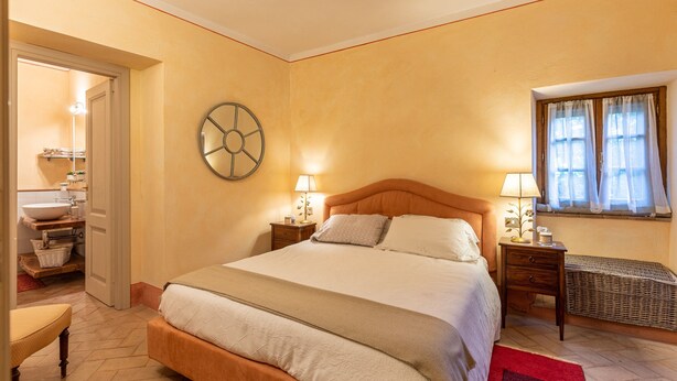 Un dormitorio con una cama grande, mesitas de noche, un reloj de pared, una ventana con cortinas y un baño con lavamanos y repisas.