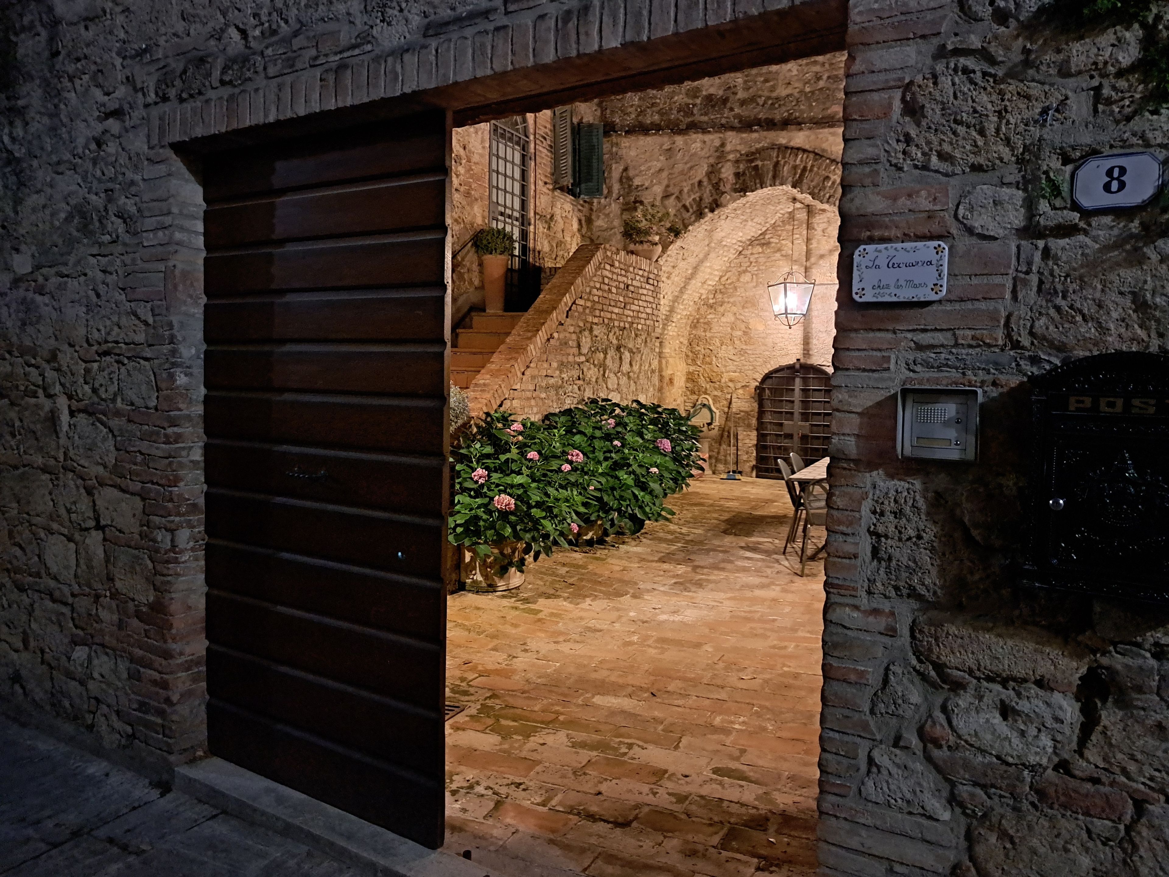 Un corridoio con pareti in pietra, una porta in legno, una scala e una pianta in vaso con fiori rosa.