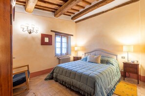 Un dormitorio con cama, cabecera de madera, dos mesitas de noche, una lámpara de pared y un radiador, además de una ventana con cortinas.