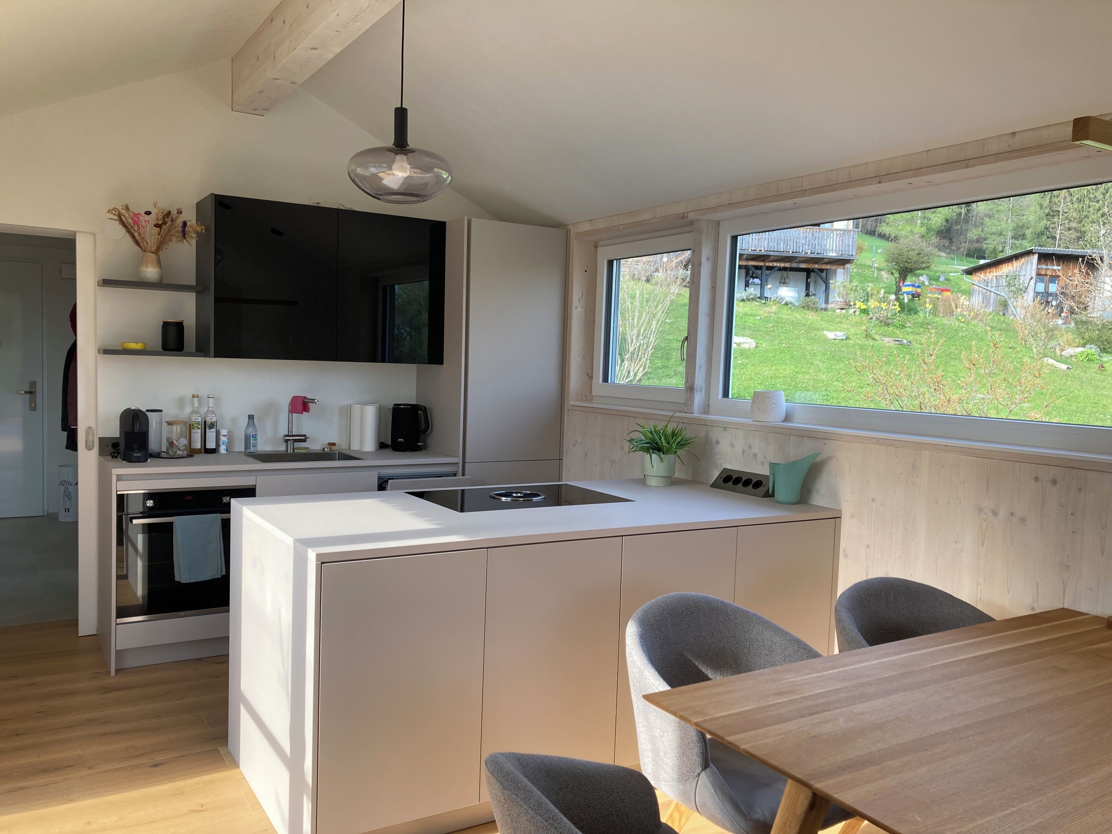 A modern kitchen with a built-in island, a dining area with a wooden table, and a window with a view of a green landscape.