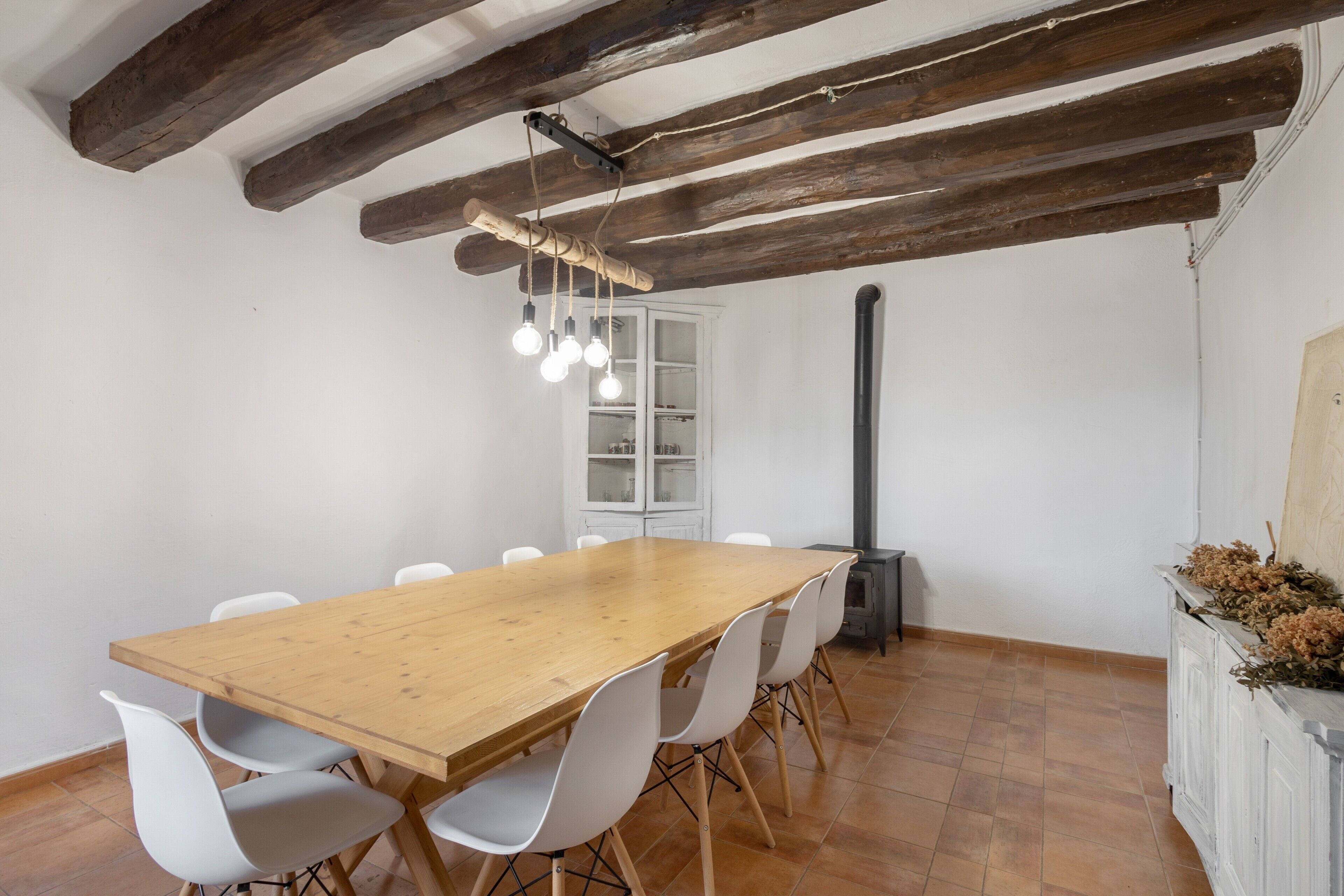 Un comedor con una mesa de madera, sillas blancas, vigas de madera a la vista, una lámpara colgante y un armario blanco con puertas de cristal.