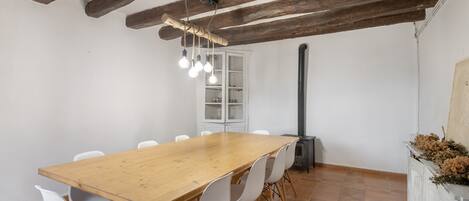 Un comedor con una mesa de madera, sillas blancas, vigas de madera a la vista, una lámpara colgante y un armario blanco con puertas de cristal.