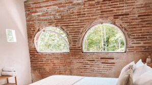 A bedroom with a brick wall, two arched windows, a bed with white linens, and a wooden chair.
