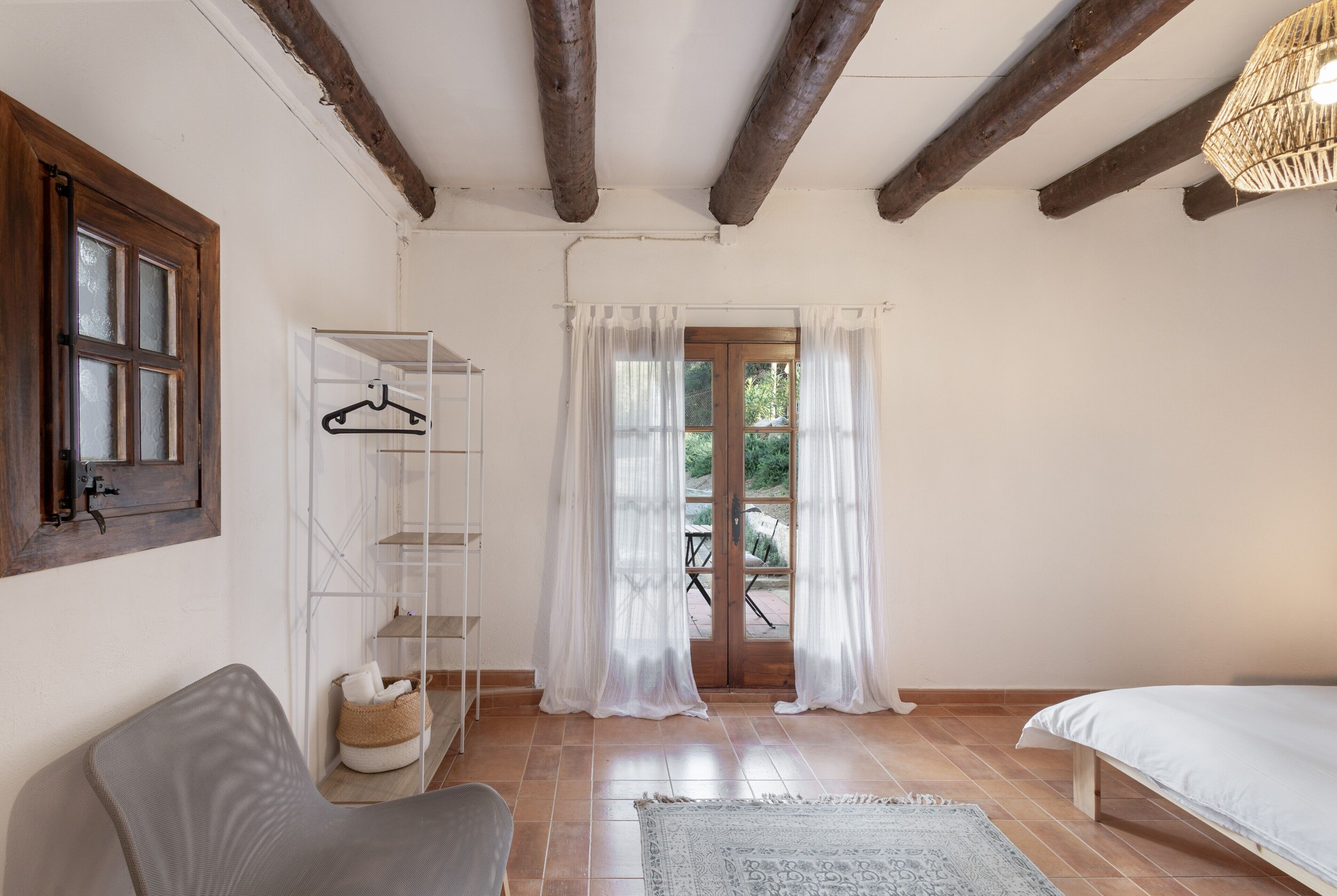 Ein Schlafzimmer mit Bett, Stuhl, Kleiderhaken und Blick ins Freie durch eine Tür.