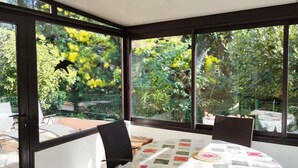 Un invernadero con una mesa y sillas, ventanas amplias con vistas a árboles y una valla.