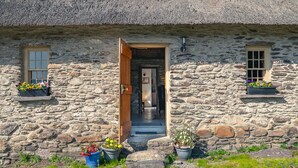 Une petite maison en pierre avec un toit de chaume, une porte en bois et des pots de fleurs placés au sol.