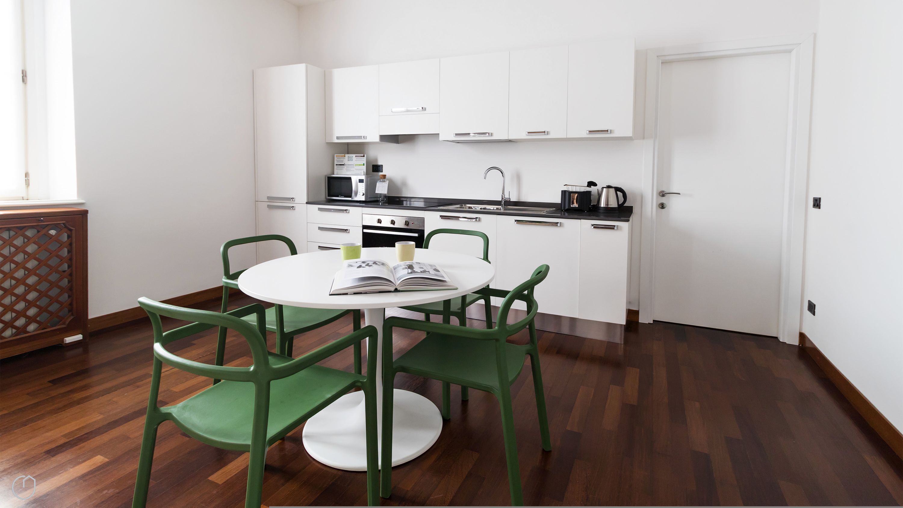 A compact kitchen with white cabinets, a black countertop, and a stainless steel sink. There is a dining table with green chairs and a book on it.