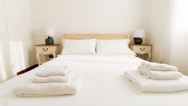 Un letto rifatto con lenzuola bianche e due cuscini, affiancato da comodini con lampade e vasi decorativi.
