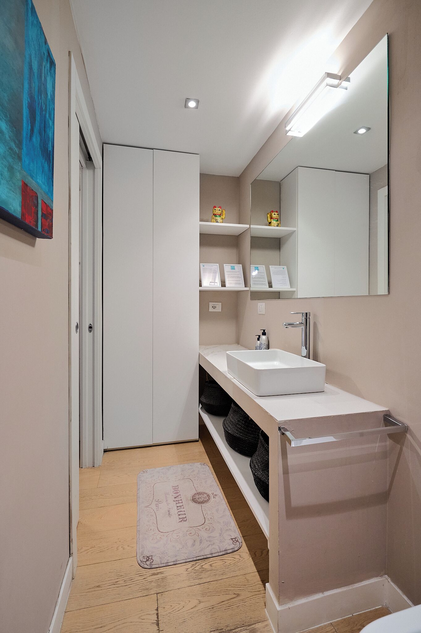 Une salle de bain moderne avec un lavabo blanc, un miroir et des armoires. On y trouve un tapis de sol en bois et une petite étagère avec des articles de toilette.