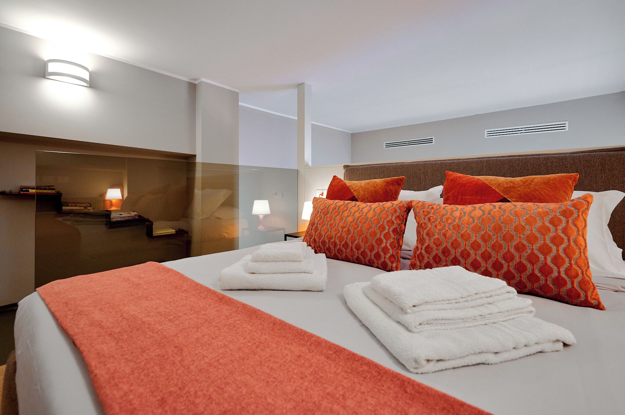 Een hotelkamer met een net opgemaakt bed, een rode deken en kussens met een oranje patroon. Er zijn twee bedlampen en een klein tafeltje met boeken en een lamp.
