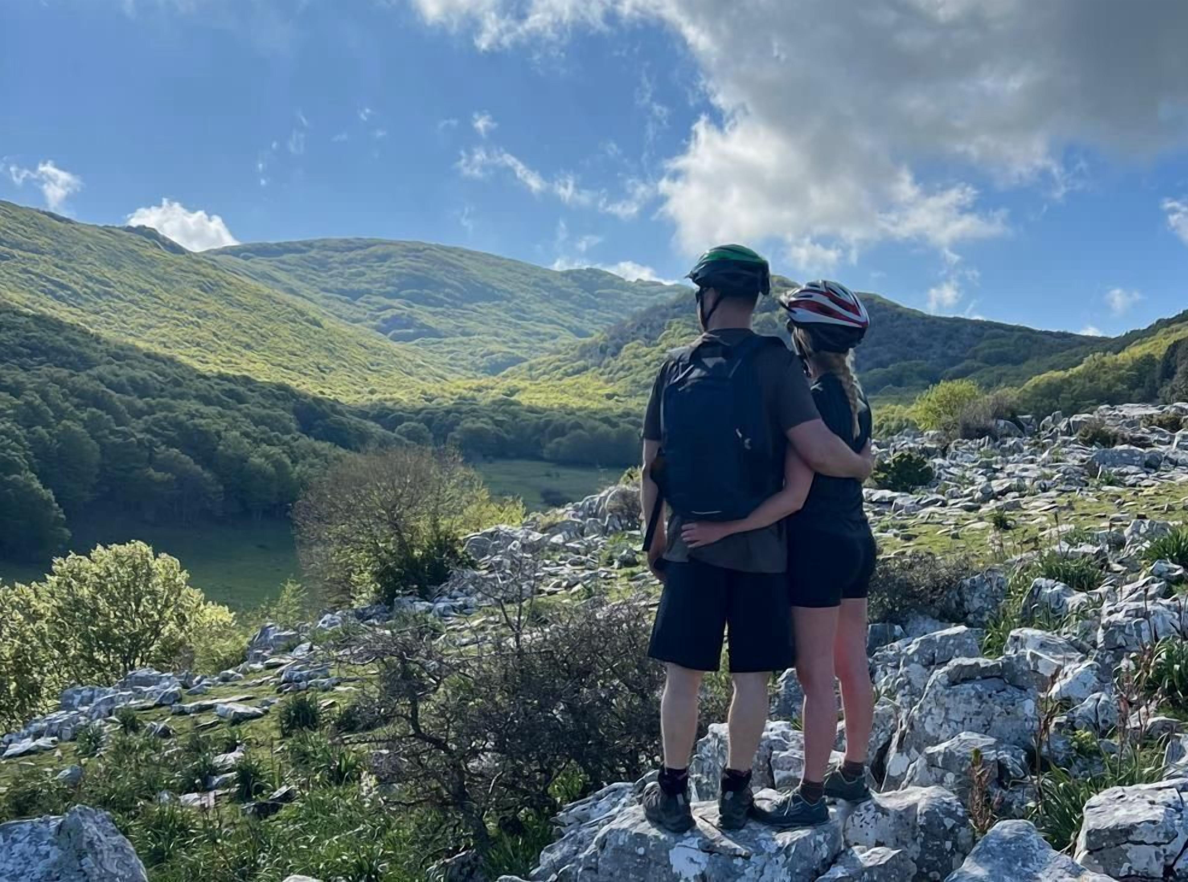 Deux personnes se tiennent debout sur un terrain rocheux, avec un paysage montagneux et un lac en arrière-plan.