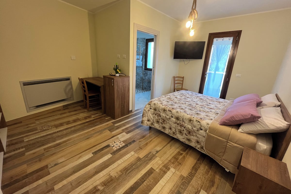 Een slaapkamer met een bed, een bureau, een stoel, een televisie en een deur naar een balkon.