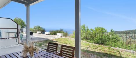 A wooden table with chairs and a vase of dried flowers, set under a covered patio with a view of the sea.