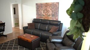 En stue med en sort lædersofa, en træpuf og et mønstret tæppe.