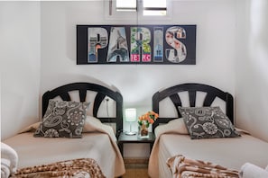 Habitación de hotel con dos camas, una mesita de noche, una lámpara y un adorno de pared con la palabra 'PARIS'.