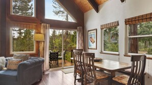A cozy room with a blue sofa, wooden dining table, and chairs. Large windows offer views of trees and a poster on the wall. - The Sunset Getaway - Hot Tub, Dog Friendly (Carnelian Bay)