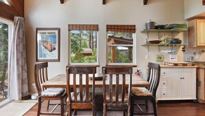 A dining area with a wooden table and chairs, a kitchen with white cabinets, and a window with a view of trees and a cabin. - The Sunset Getaway - Hot Tub, Dog Friendly (Carnelian Bay)