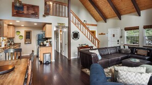 A spacious living area with a high ceiling and wooden beams, a dining area with a kitchen in the background, and a staircase leading to an upper level. - The Sunset Getaway - Hot Tub, Dog Friendly (Carnelian Bay)