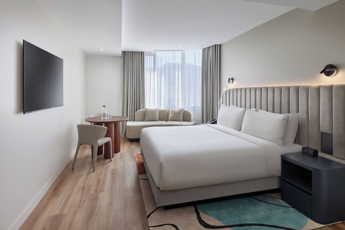 Une chambre d’hôtel moderne avec un grand lit, une table de chevet avec une horloge, une petite table ronde avec des chaises et un téléviseur fixé au mur.