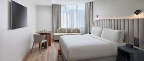 Una habitación de hotel moderna con una cama grande, una mesita de noche con un reloj, una pequeña mesa redonda con sillas y un televisor montado en la pared.