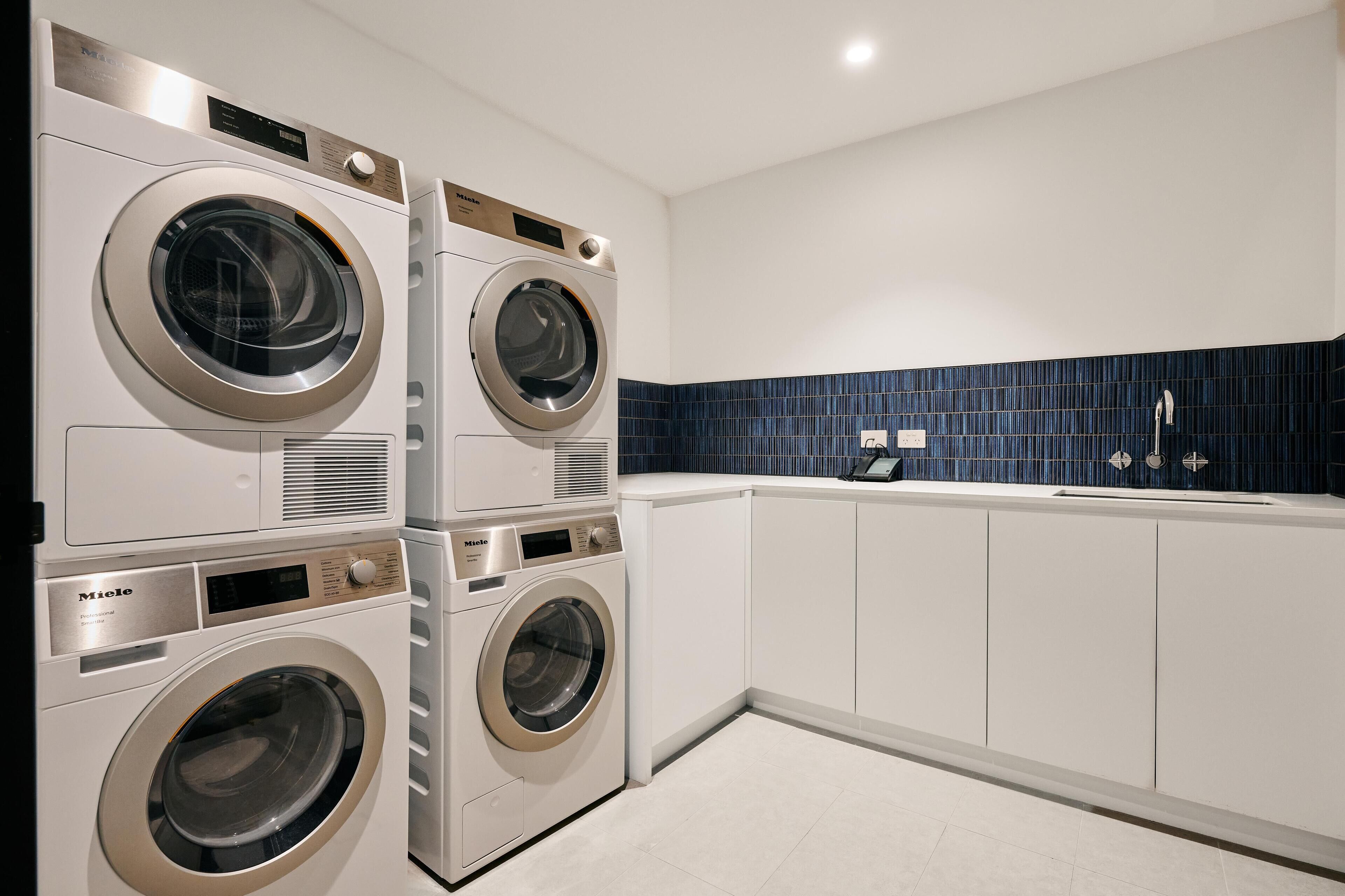 A modern laundry room with stacked front-loading washing machines, a tiled backsplash, and a built-in sink.