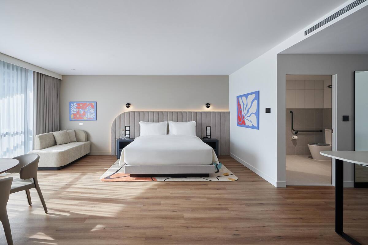 Une chambre d’hôtel moderne, dotée d’un grand lit, d’un canapé, d’une table à manger et d’une salle de bain visible à travers une porte ouverte.