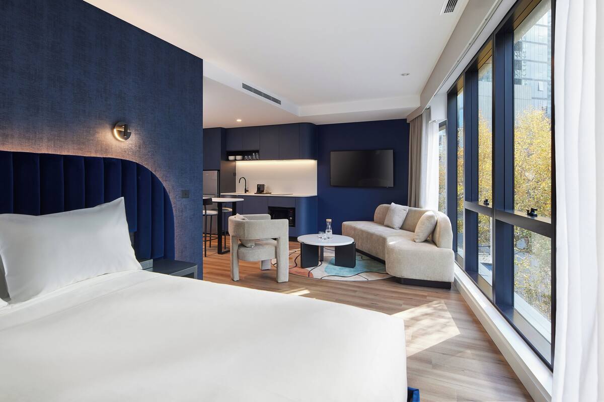 Une chambre d’hôtel moderne dotée d’un grand lit, d’un coin salon avec un canapé et des fauteuils, d’une petite table et d’un téléviseur fixé au mur.