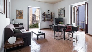 A modern living room with a sofa, coffee table, and dining area. There is a bookshelf with books and decorative items, a TV, and a balcony with a view of buildings. - Casa all Anfiteatro di Catania by Wonderful Italy (Catania)