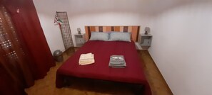 A bedroom with a wooden headboard, two bedside tables with lamps, a red bedspread, and a pair of folded towels.