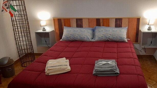 Una cama con colcha roja, dos almohadas, dos lámparas de noche, cabecera de madera y un cubo de basura pequeño.